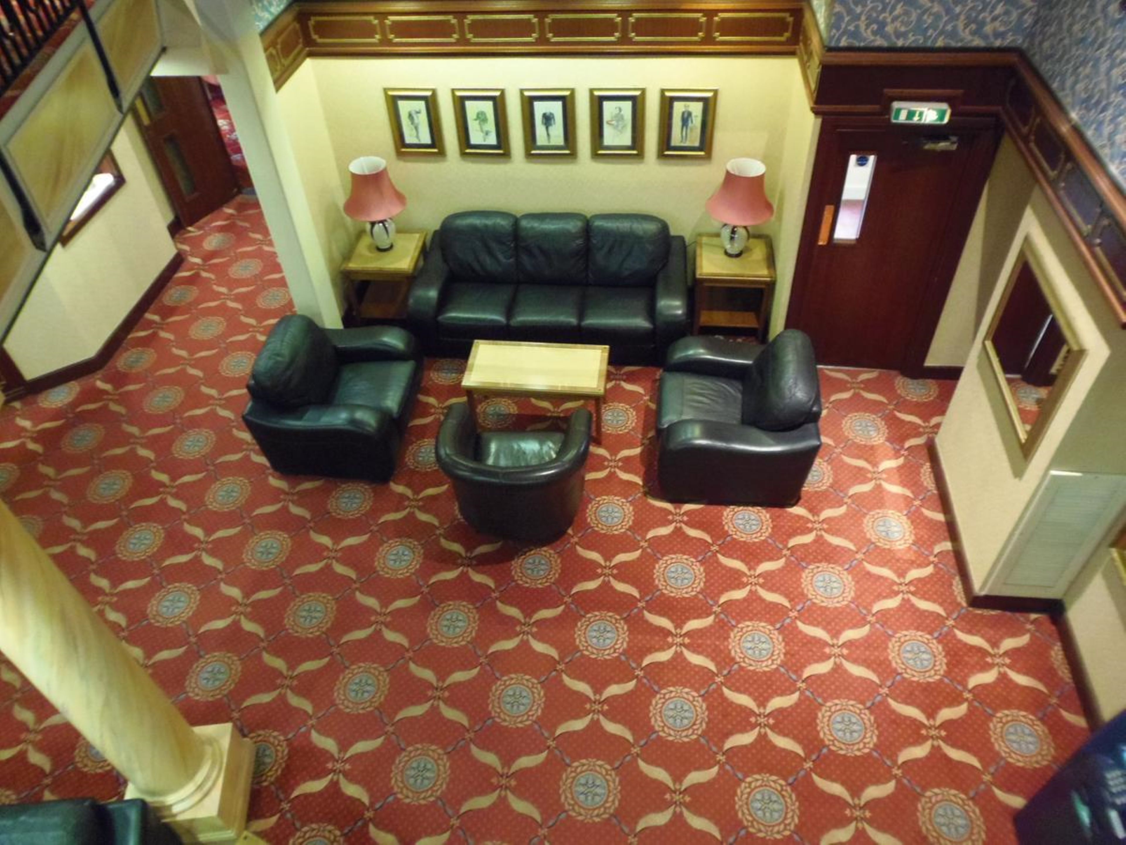A hotel lobby with a red patterned carpet, a black leather sofa set, two armchairs, a small wooden coffee table, and framed artwork on the wall.