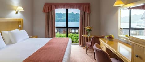 A hotel room with a large bed, a desk with a chair, a window with a view of greenery and a body of water, and a lamp on the wall.