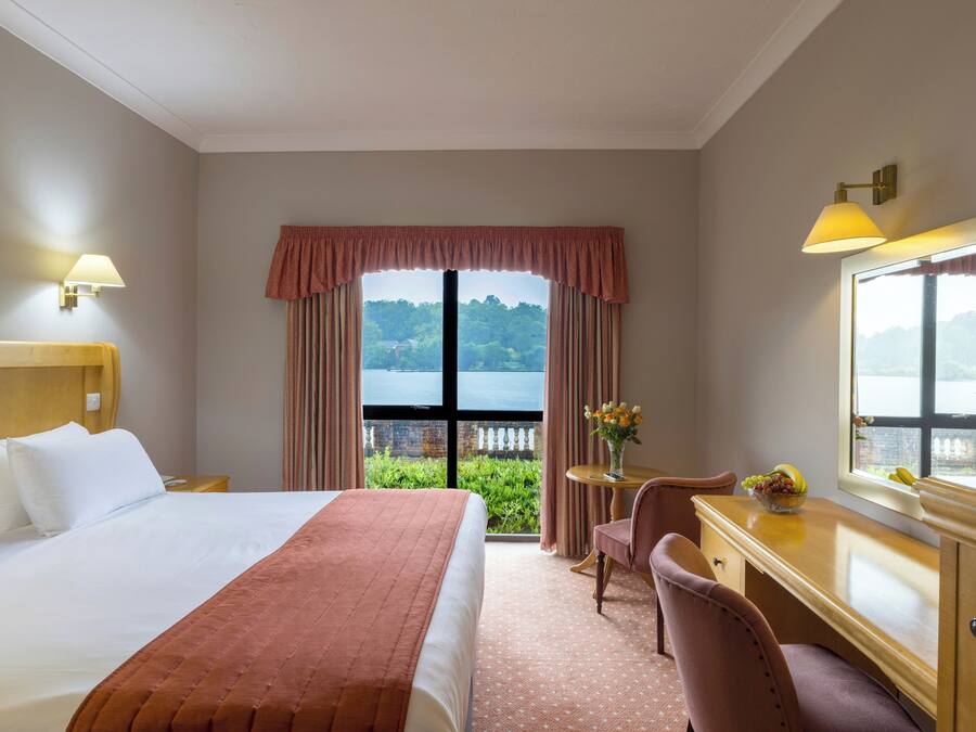 Ein Hotelzimmer mit einem großen Bett, einem Schreibtisch mit Stuhl, einem Fenster mit Blick auf Grünflächen und ein Gewässer sowie einer Wandleuchte.