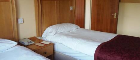 A hotel room with two single beds, wooden headboards, a nightstand with a phone and a book, and a door leading to another room.