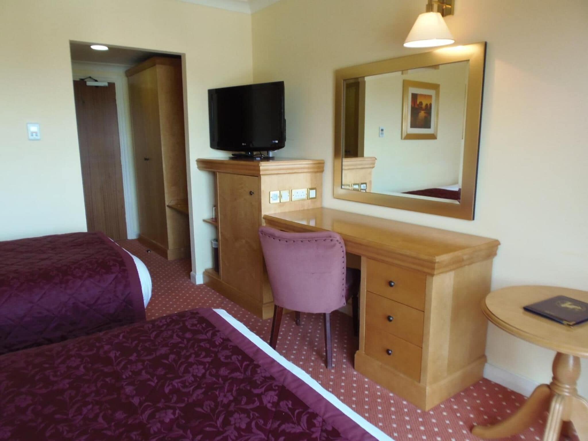 A hotel room with two beds, a wooden desk with a chair, a television, a mirror, and a small round table with a book.