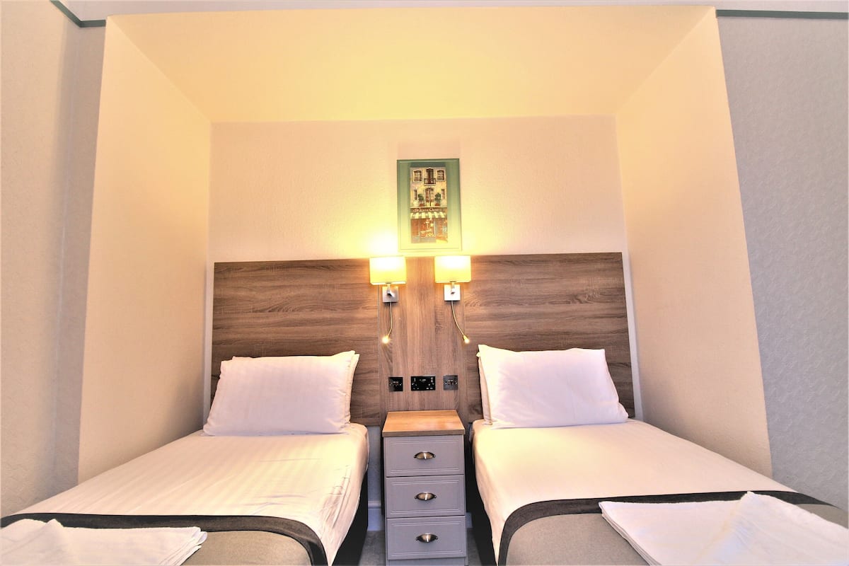 A hotel room with two beds, a wooden headboard, bedside lamps, and a small table with drawers.