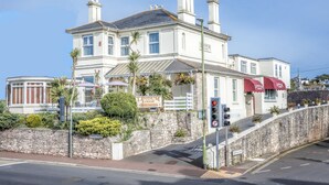 A two-story building with a sign reading "Lindum Lodge." It features a stone wall, a small garden with plants, and a red awning over the entrance. - Lindum Lodge (Torquay)