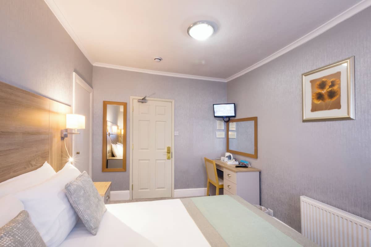 A hotel room with a bed, bedside table, lamp, desk, chair, TV, and a door.