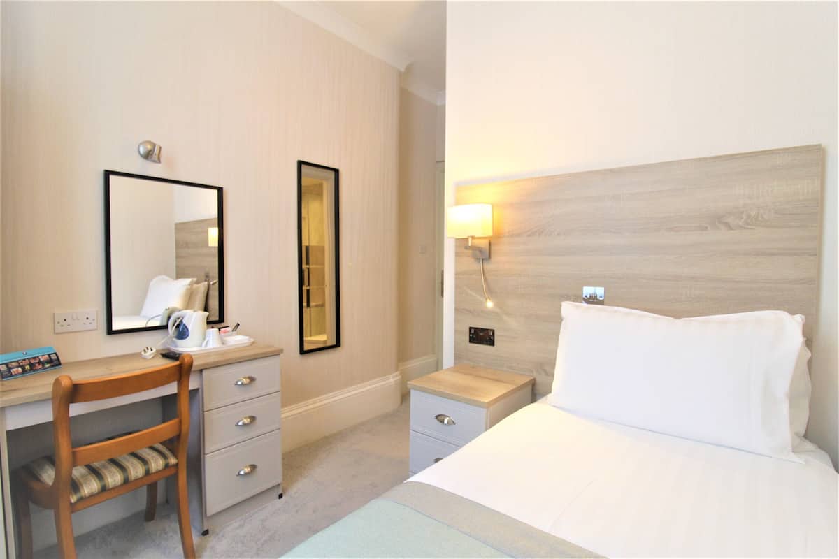 A hotel room with a wooden desk, a chair, a bed with white bedding, a nightstand, a lamp, and a mirror.