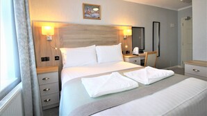 A hotel room with a large bed, two bedside tables, a nightstand, a mirror, and a window with curtains. - Lindum Lodge (Torquay)