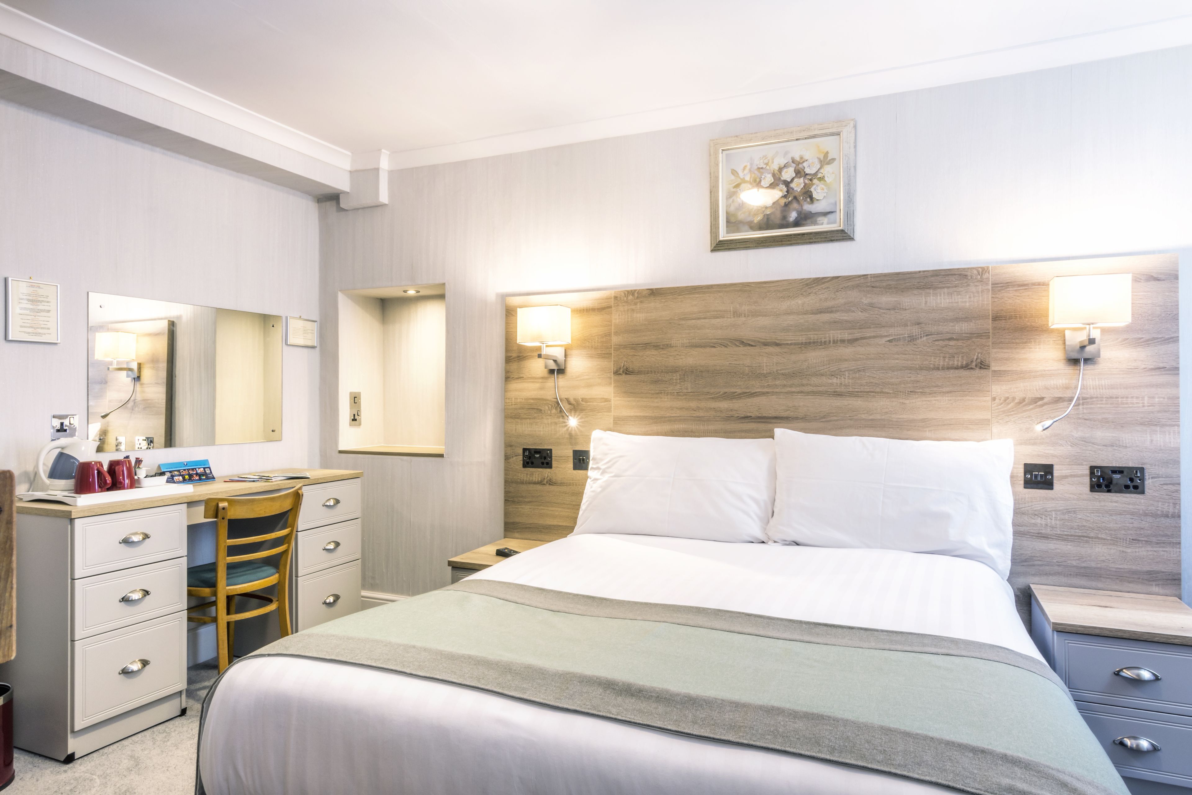 Une chambre d’hôtel comprenant un lit, un bureau avec une chaise, une table de chevet, un miroir et une lampe fixée au mur.