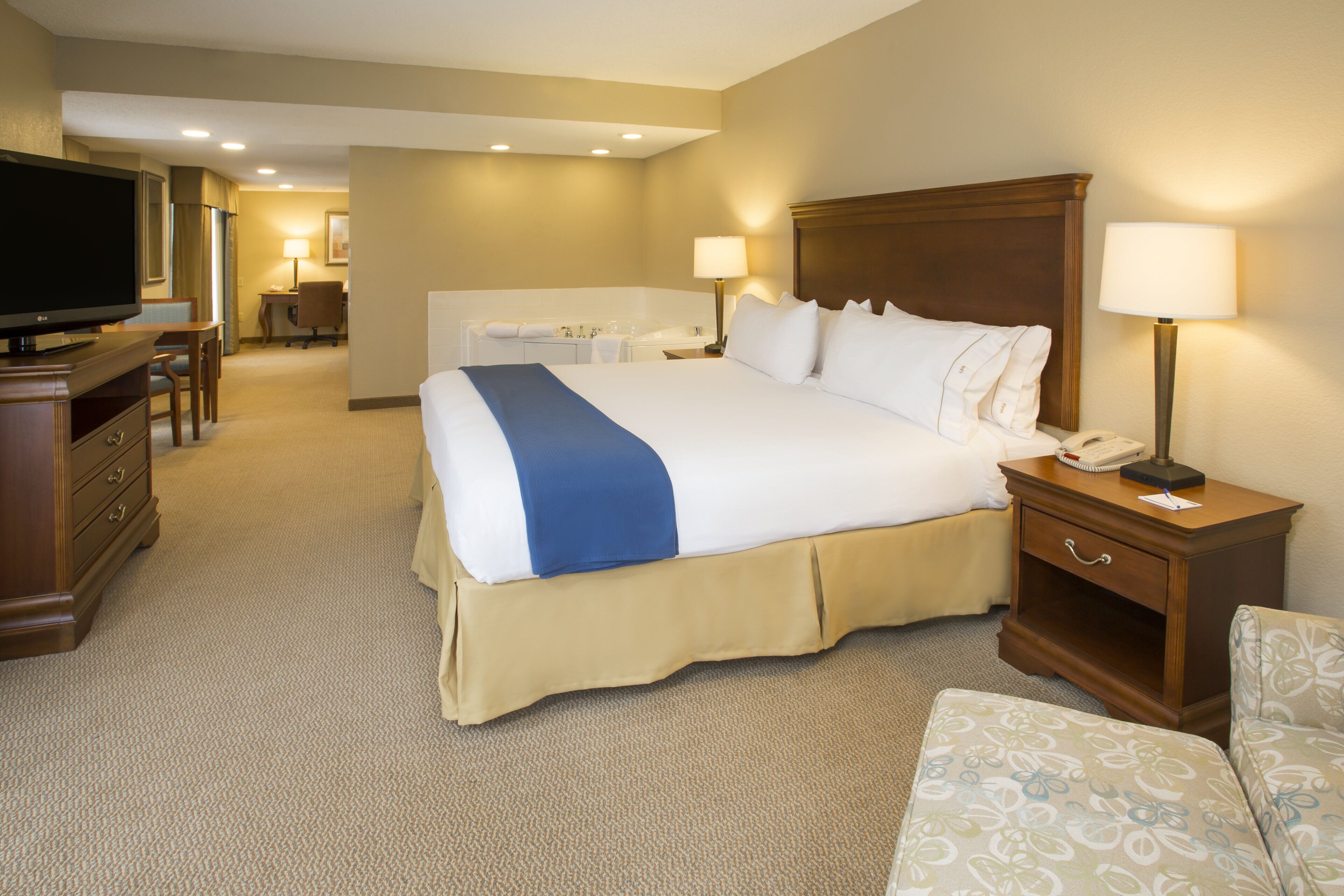 A hotel room with a large bed, a desk, a chair, a television, and a bathroom in the background.