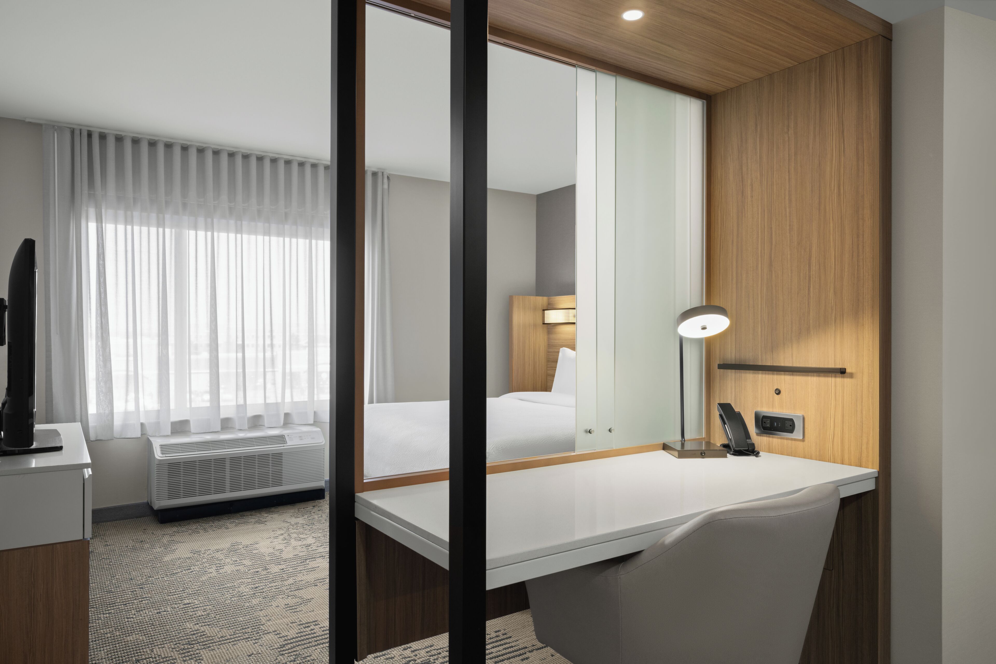 A hotel room with a bed, a desk, a chair, a lamp, a TV, and a window with curtains.