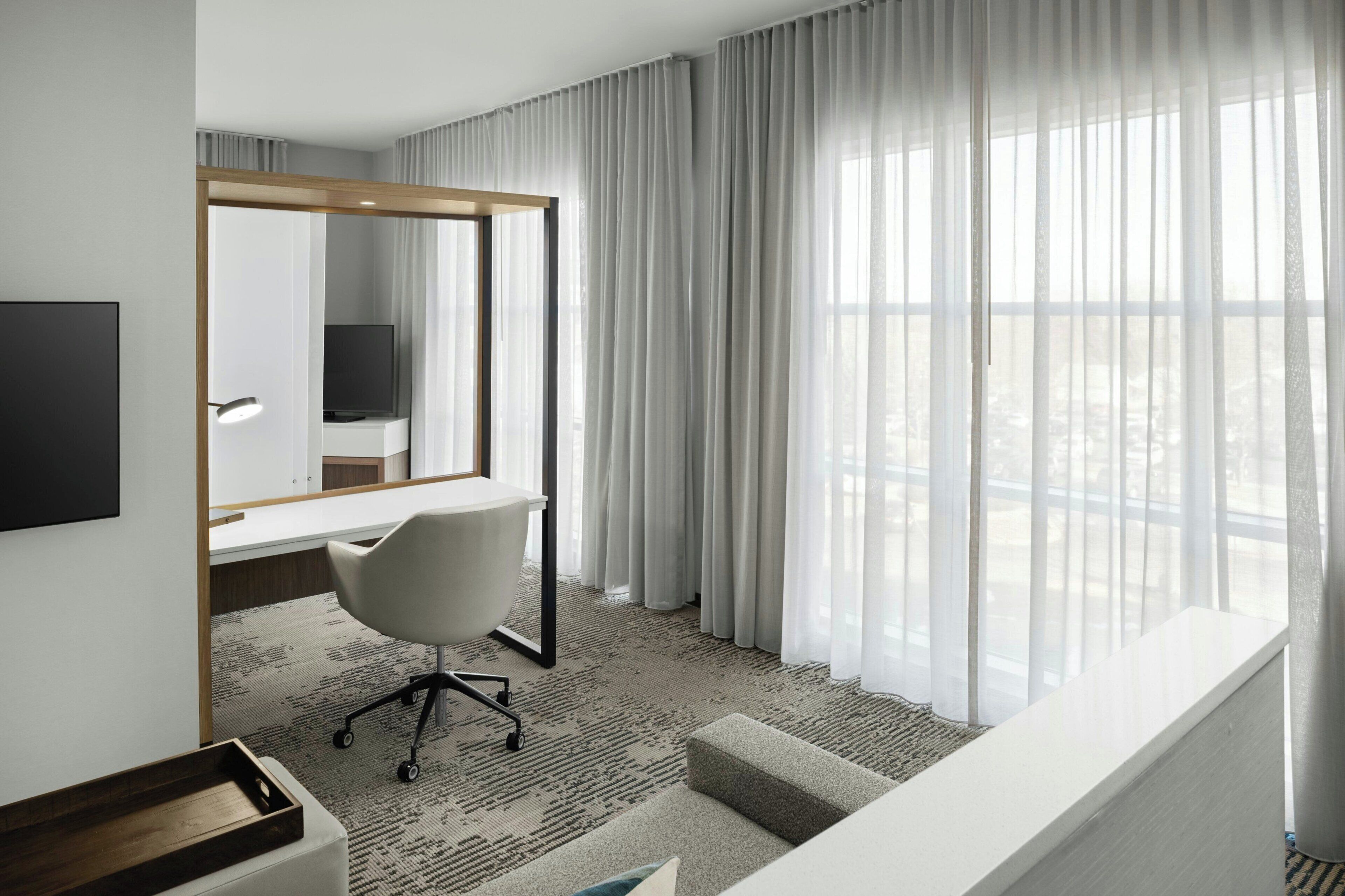 A modern hotel room with a large window, a desk with a chair, a television, and a mirror.