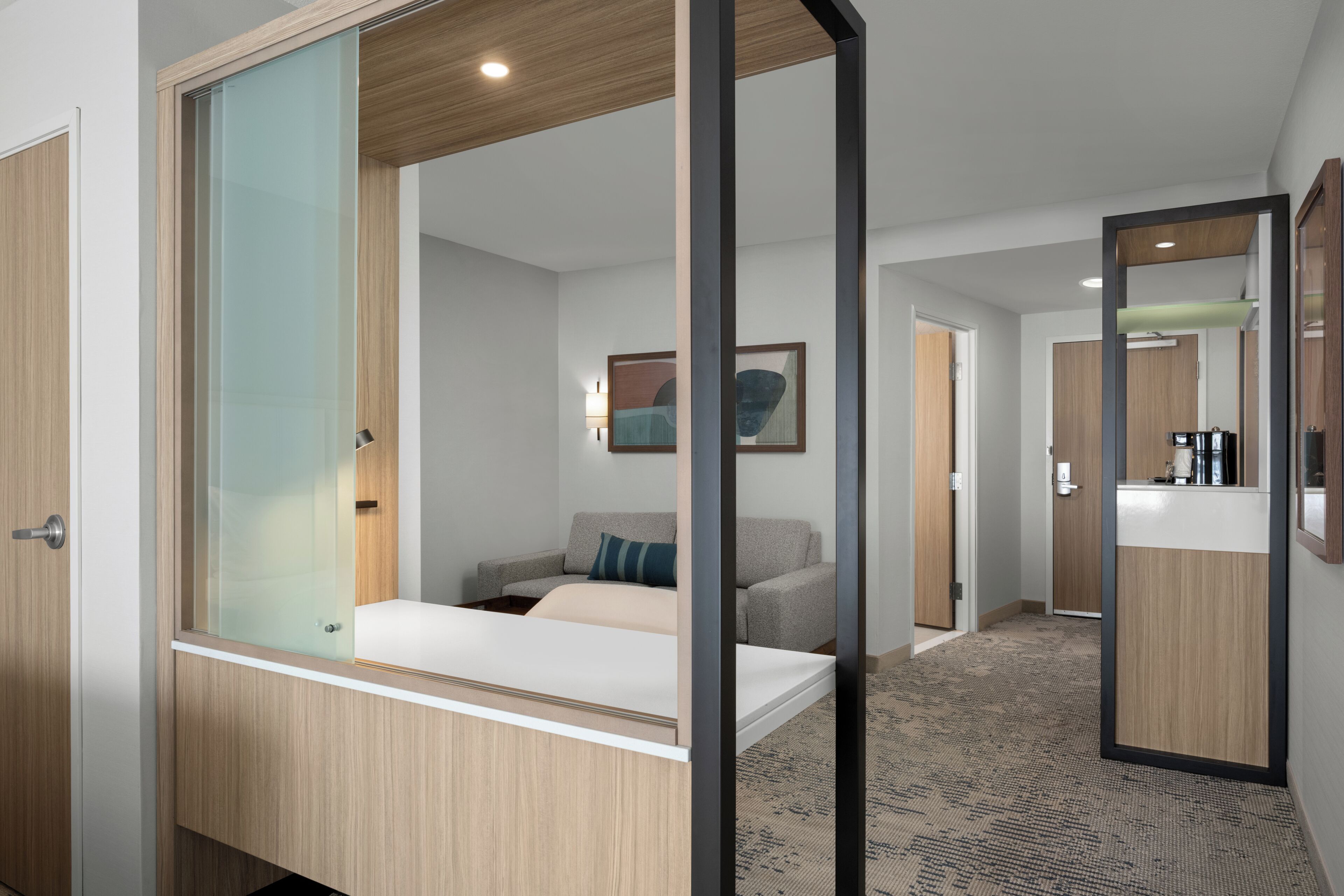 Quarto de hotel com cama, sofá, divisória de vidro, armário de madeira, espelho e corredor que dá para um banheiro.