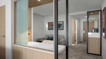 Quarto de hotel com cama, sofá, divisória de vidro, armário de madeira, espelho e corredor que dá para um banheiro.