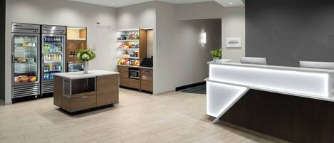 A modern hotel lobby with a reception desk, a seating area, and a refrigerated display case.