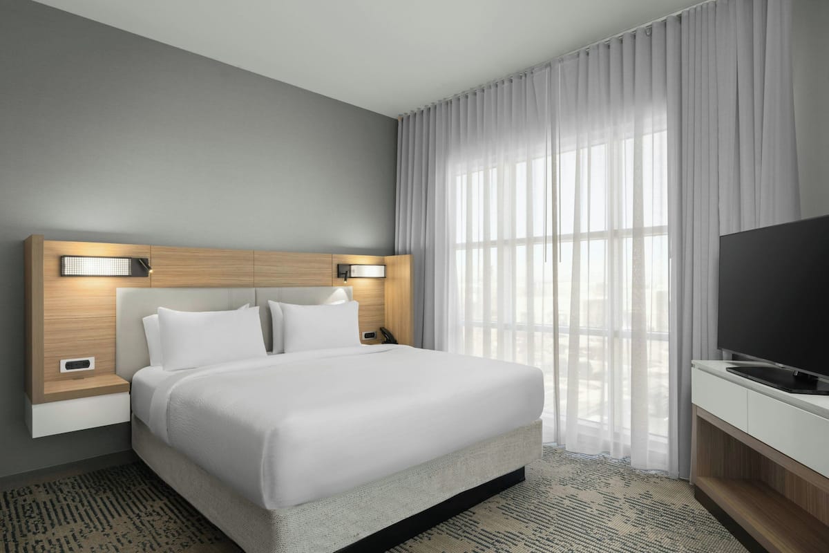 A modern hotel room with a large bed, a flat-screen TV, a bedside table, and a window with sheer curtains.
