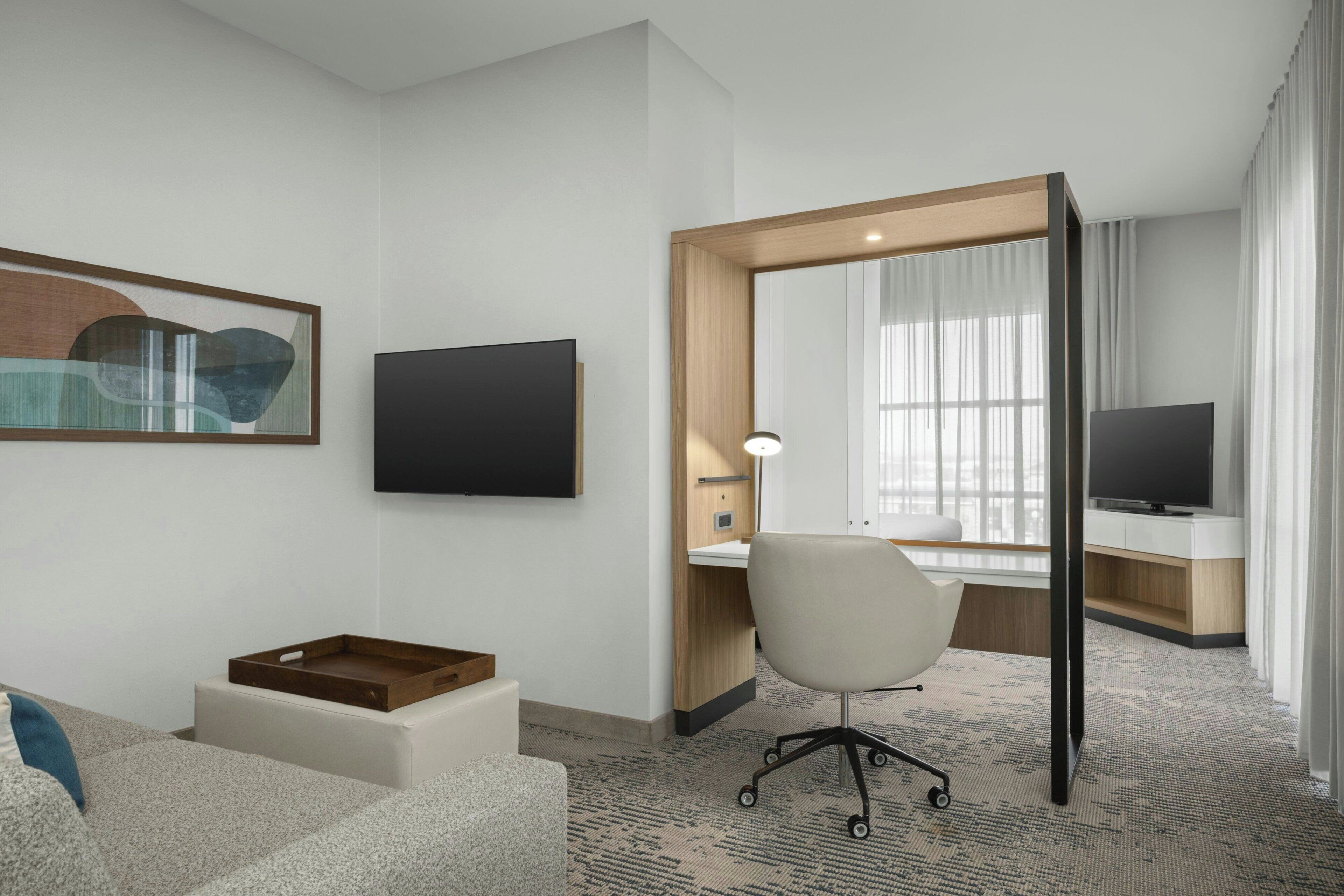 A hotel room with a desk, chair, TV, artwork, and a window with curtains.