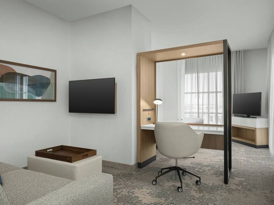 A hotel room with a desk, chair, TV, artwork, and a window with curtains.