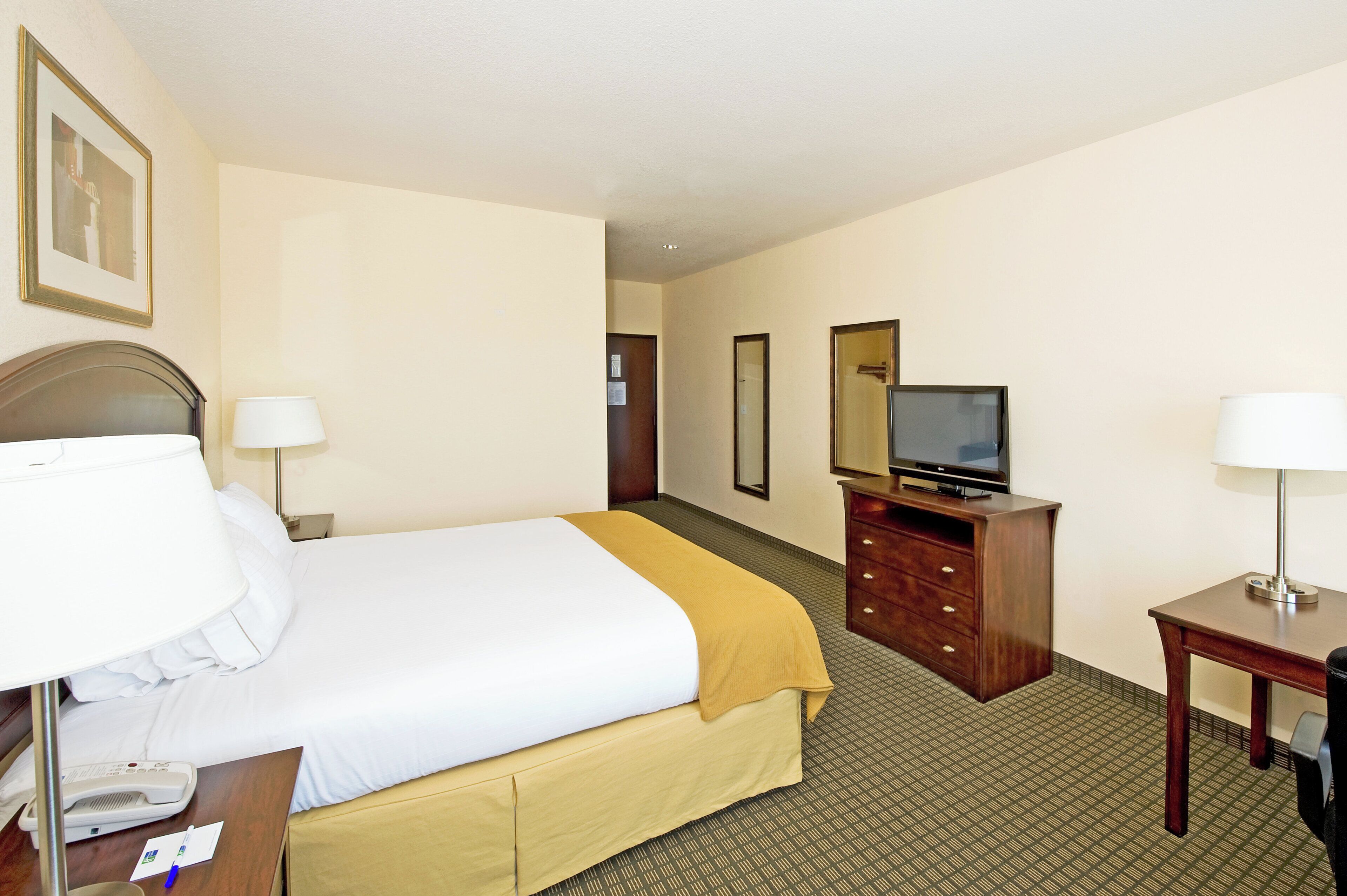 A hotel room with a bed, bedside lamp, a desk with a lamp, a television, and a dresser.