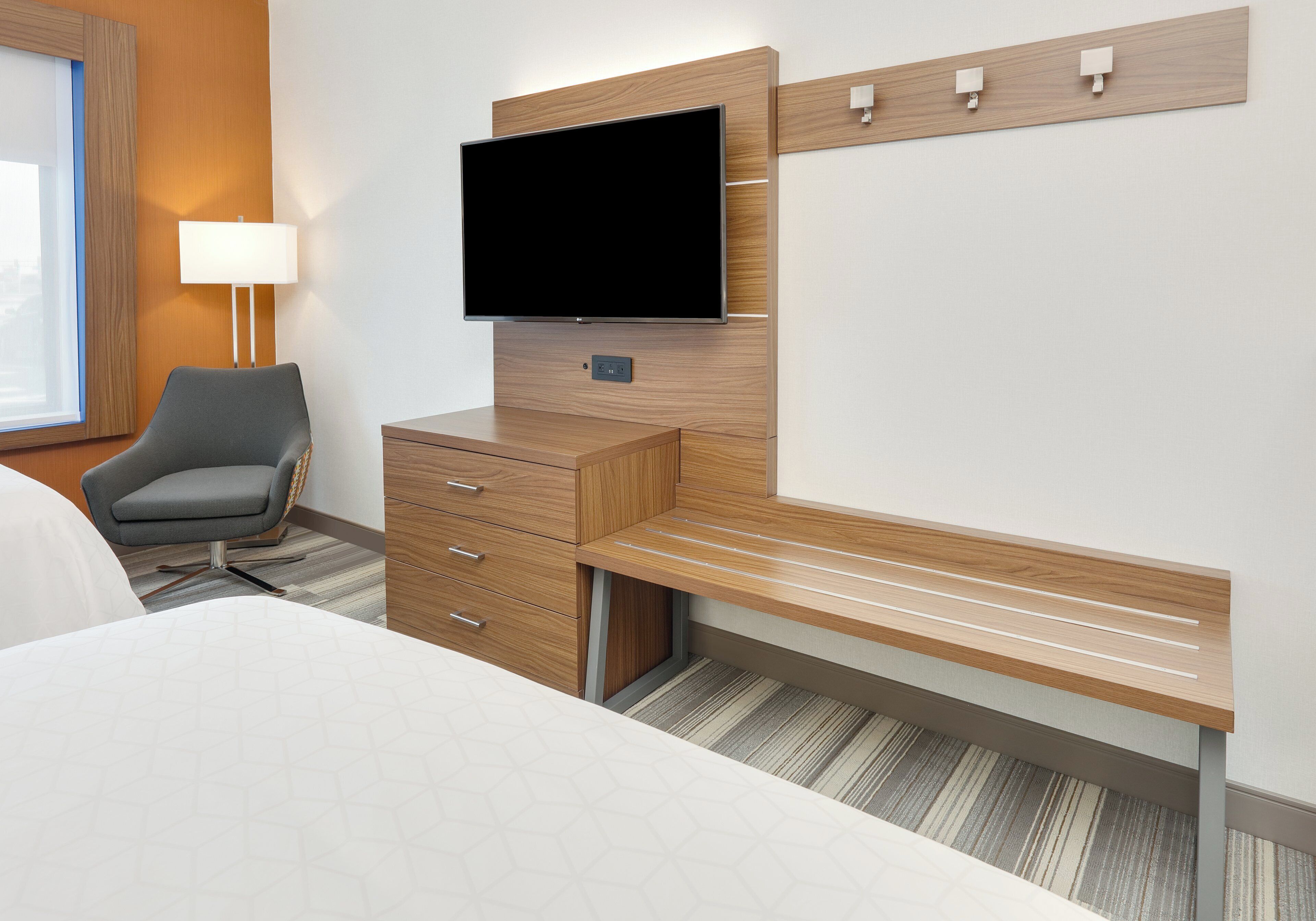 A hotel room with a bed, a desk with drawers, a chair, a flat-screen TV mounted on the wall, and a lamp.