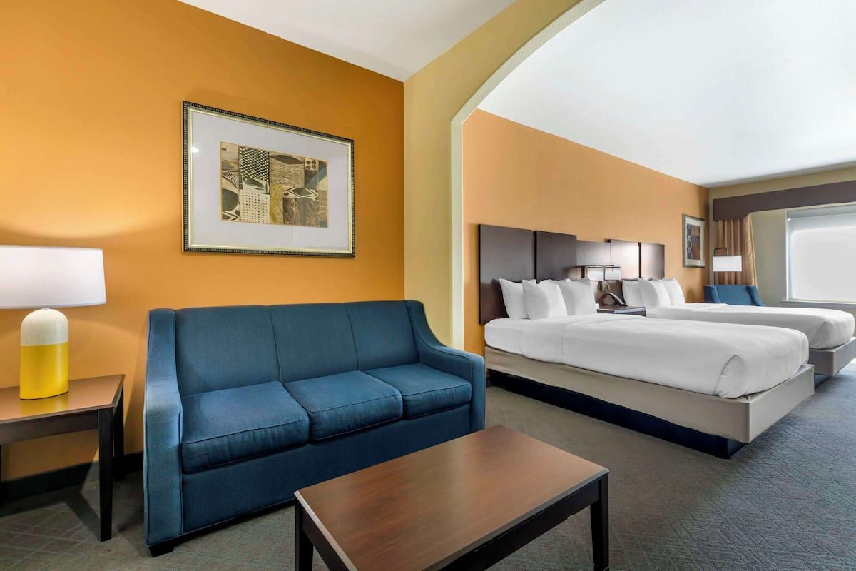 A hotel room with a blue sofa, a bed with white bedding, a bedside table with a lamp, and a framed picture on the wall.