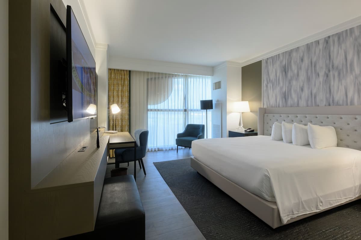 A hotel room with a large bed, a desk with a chair, a flat-screen TV mounted on the wall, and a window with curtains.
