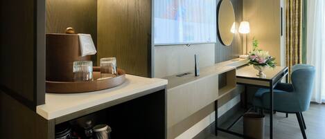 Habitación de hotel moderna con tocador de madera, un escritorio con silla, un espejo grande y una pantalla de proyector mostrando el nombre del hotel.