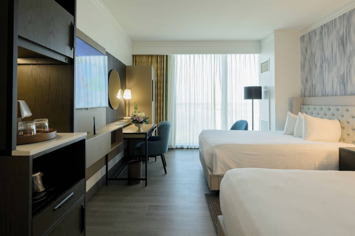 A hotel room with two beds, a desk with a chair, a mirror, and a lamp.