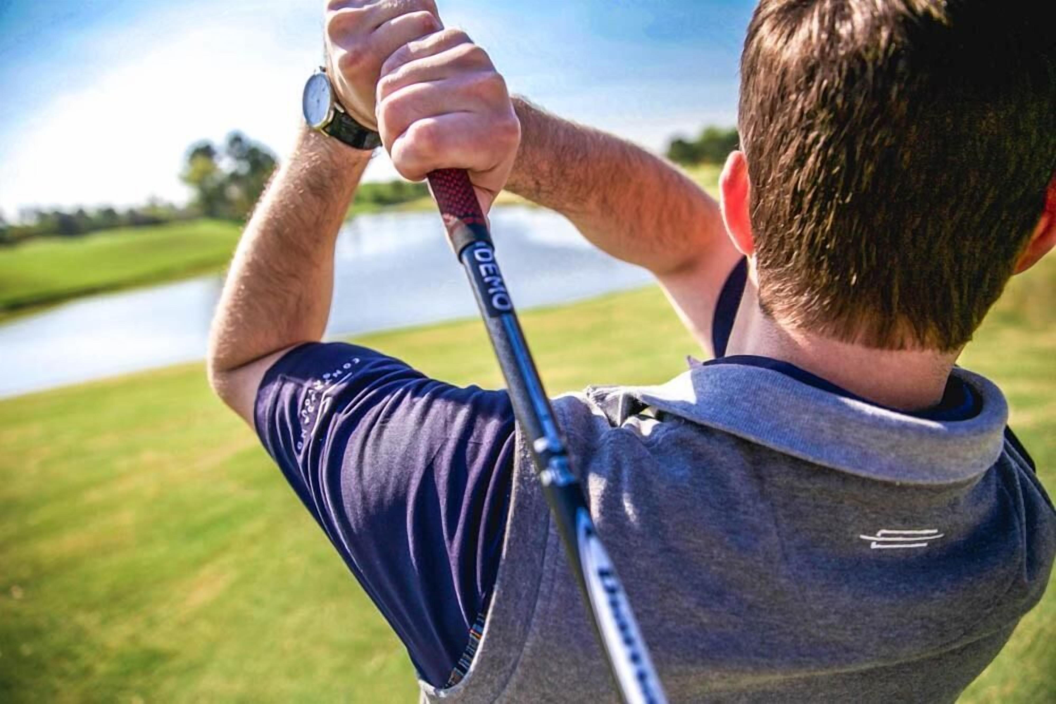 En person som håller i en golfklubba och har en klocka på handleden, och som står på en golfbana med en vattenyta i bakgrunden.