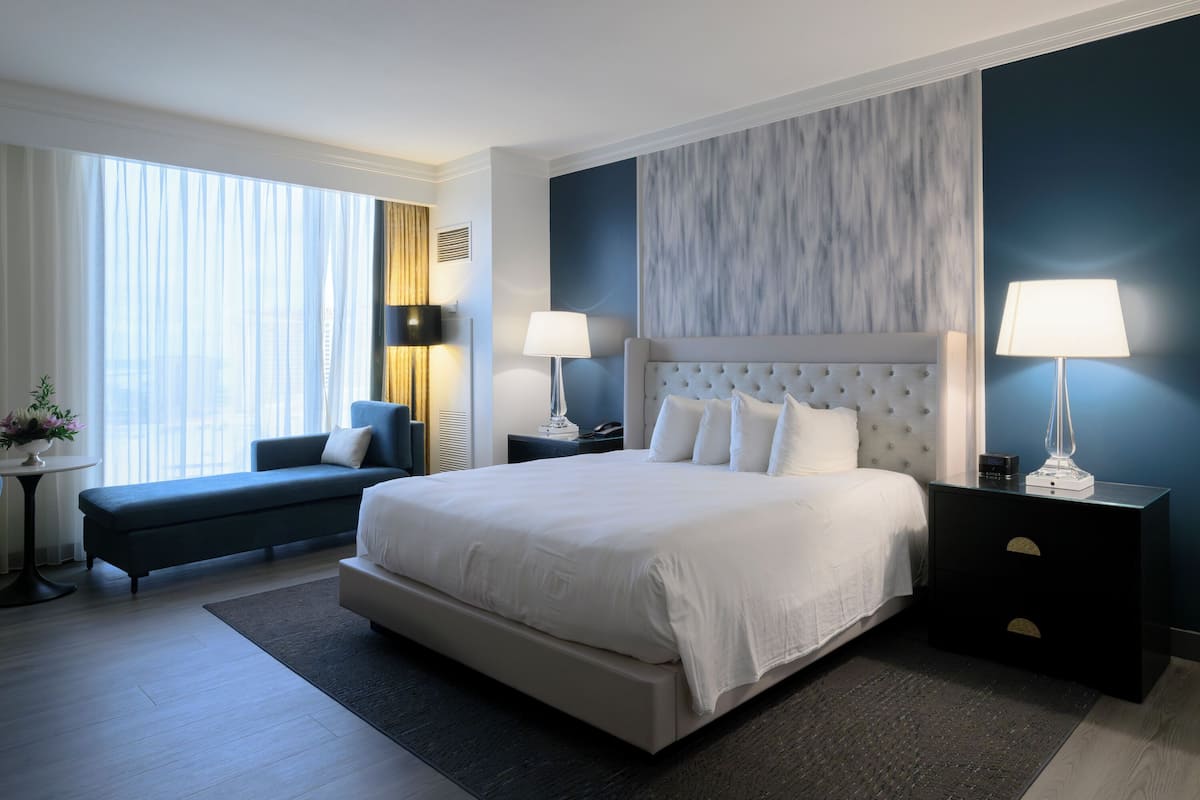 A modern hotel room with a large bed, a sofa, two bedside tables with lamps, and a view of the city through large windows.