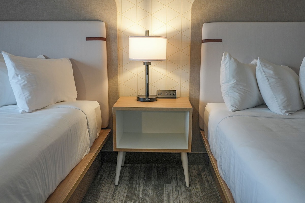 A hotel room with two beds, a nightstand with a lamp, and a control panel.