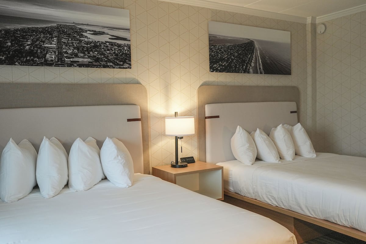 A hotel room with two beds, a nightstand with a lamp, and two wall-mounted paintings.