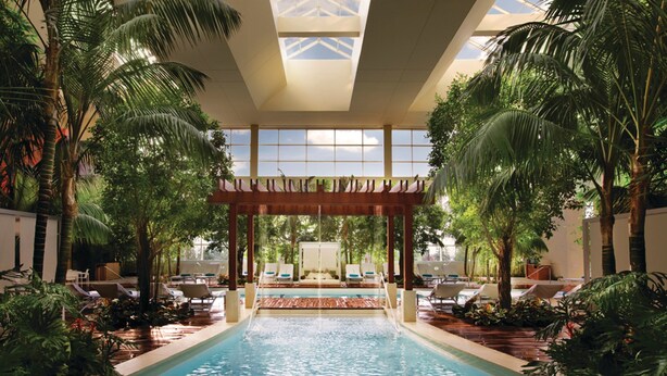 An indoor swimming pool with lounge chairs and palm trees, surrounded by a covered area with a view of the sky. - MGM Tower at Borgata (Atlantic City)