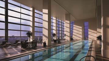 A modern indoor swimming pool with large windows, lounge chairs, and a view of the cityscape.