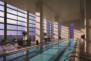 A modern indoor swimming pool with large windows, lounge chairs, and a view of the cityscape.