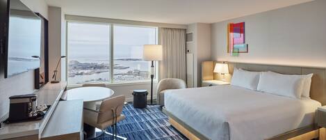 A hotel room with a bed, a desk, a chair, a lamp, and a view of the city through large windows.