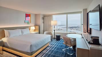 A hotel room with a large bed, a desk, a chair, a TV, and a view of the city.