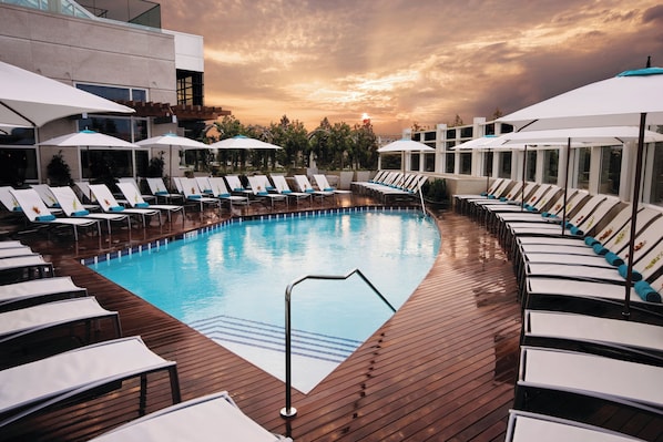 A swimming pool with lounge chairs and umbrellas, surrounded by a wooden deck and glass walls.
