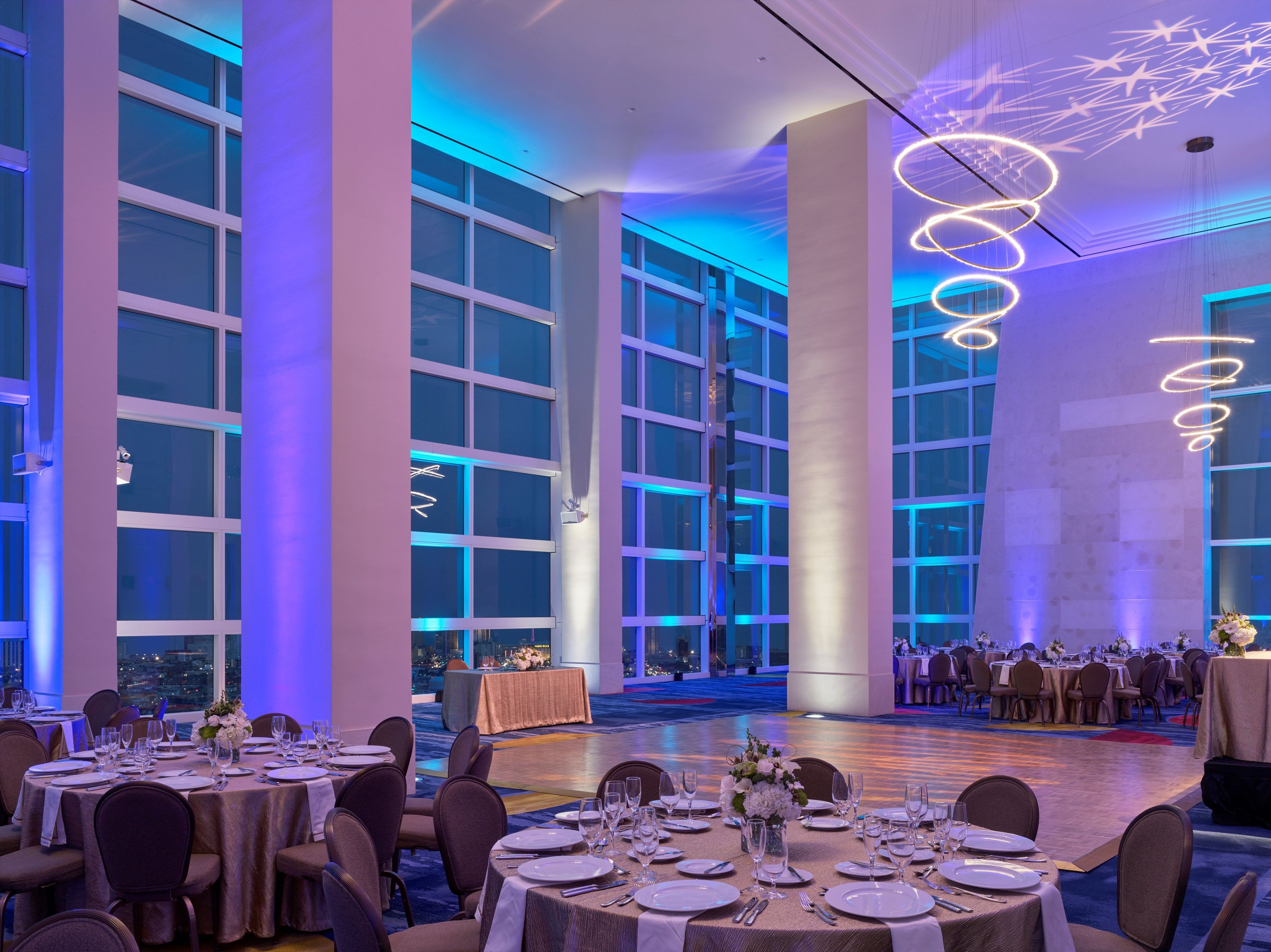 A banquet hall with round tables set for dining, large windows, and modern lighting fixtures.