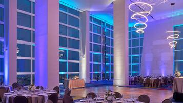 A banquet hall with round tables set for dining, large windows, and modern lighting fixtures.