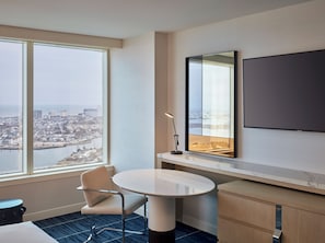 A hotel room with a large window, a round table, a chair, a desk, and a mirror.