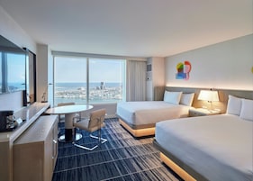 Habitación de hotel con dos camas, un escritorio, una silla, un televisor y un amplio ventanal con vistas a la ciudad.