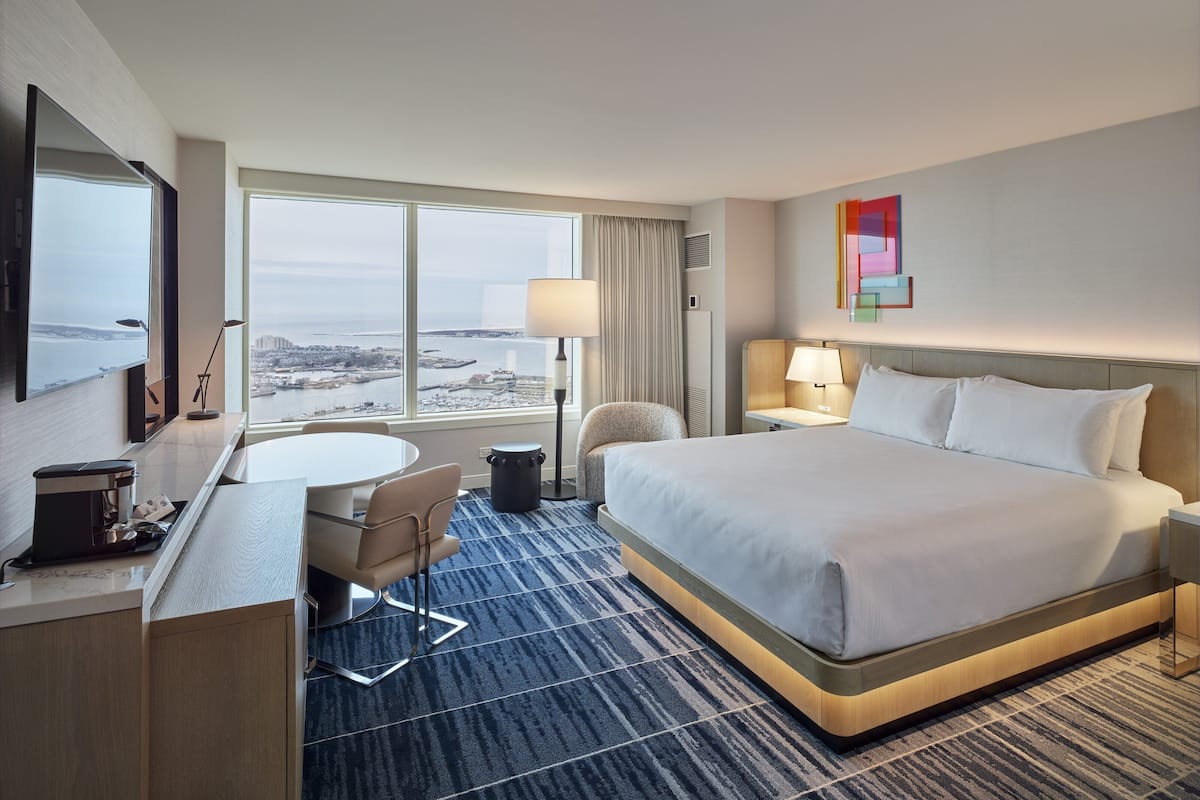 Une chambre d’hôtel avec un grand lit, un bureau avec une chaise, une petite table et une vue sur la ville grâce à de grandes fenêtres.