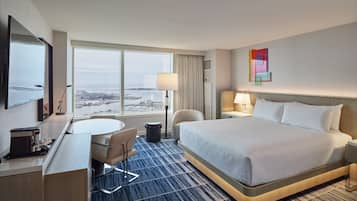 A hotel room with a large bed, a desk with a chair, a small table, and a view of the city through large windows.