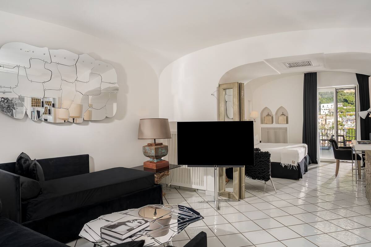 A modern hotel room with a black sofa, a flat-screen TV, a dining area with a table and chairs, and a large mirror on the wall.