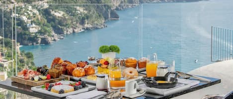 A breakfast table with a variety of pastries, fruits, and beverages, overlooking a coastal landscape with buildings and cliffs.