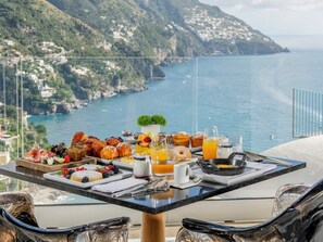 Una mesa de desayuno con diversos pasteles, frutas y bebidas, con vistas a un paisaje costero con edificios y acantilados.