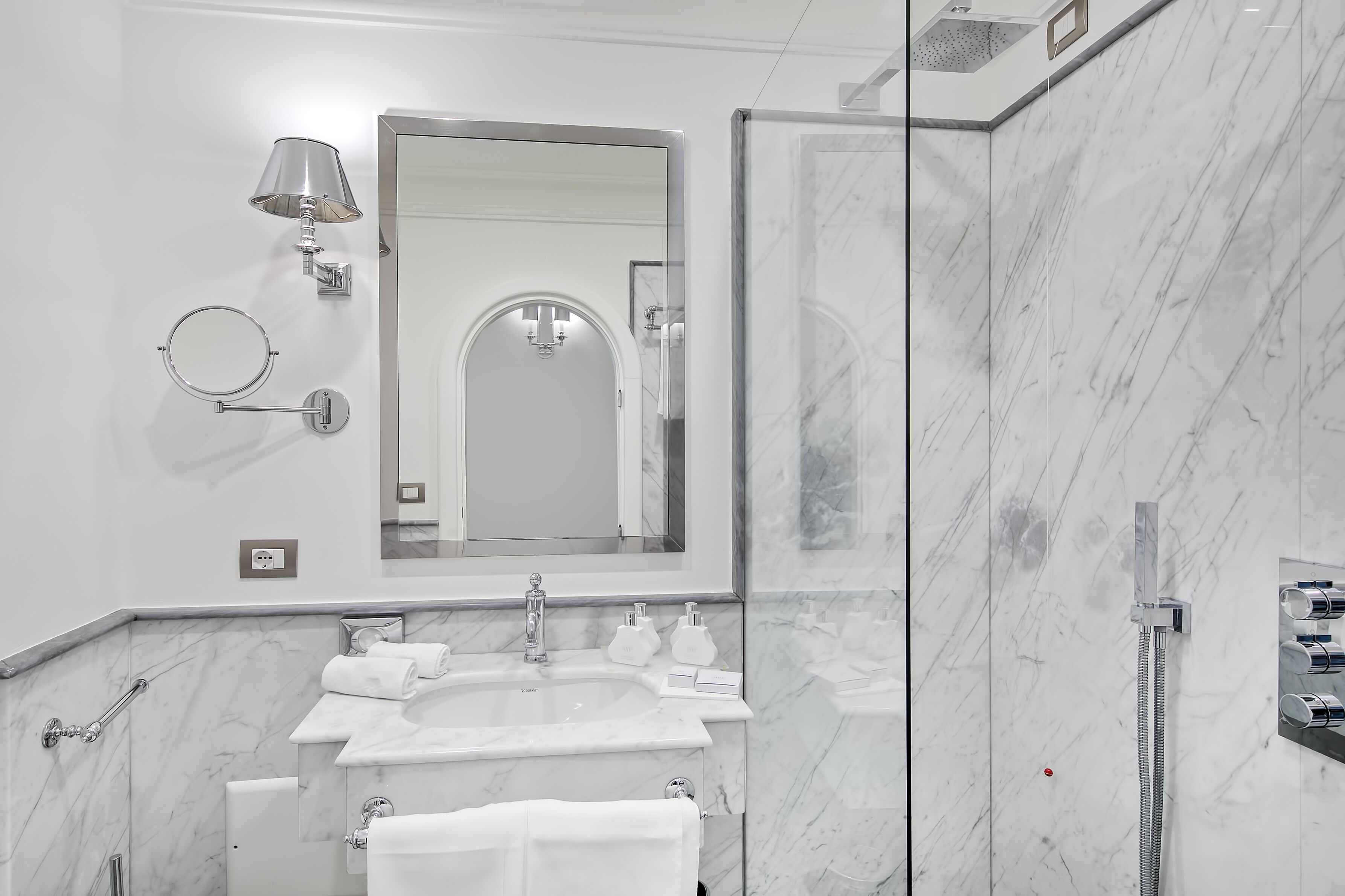 Un baño moderno con lavamanos de mármol, un espejo grande, un cabezal de ducha de mano y un perchero.