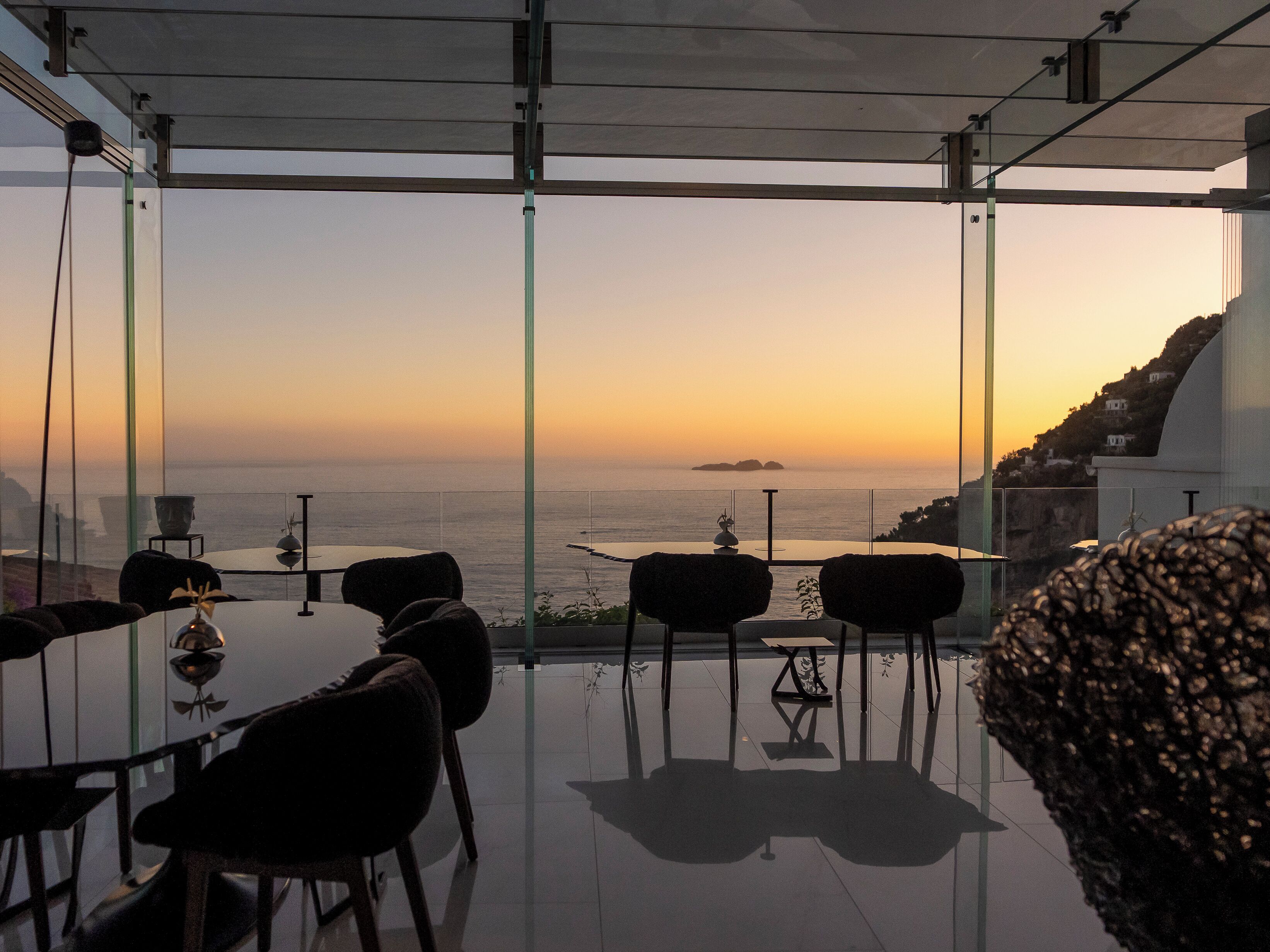Un espace repas moderne avec une table et des chaises en verre, offrant une vue sur la côte au coucher du soleil.
