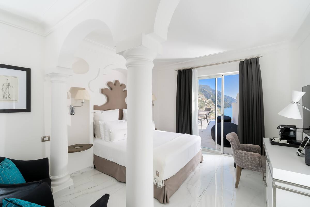 A hotel room with a large bed, a balcony with a view of the mountains, and a desk with a lamp.