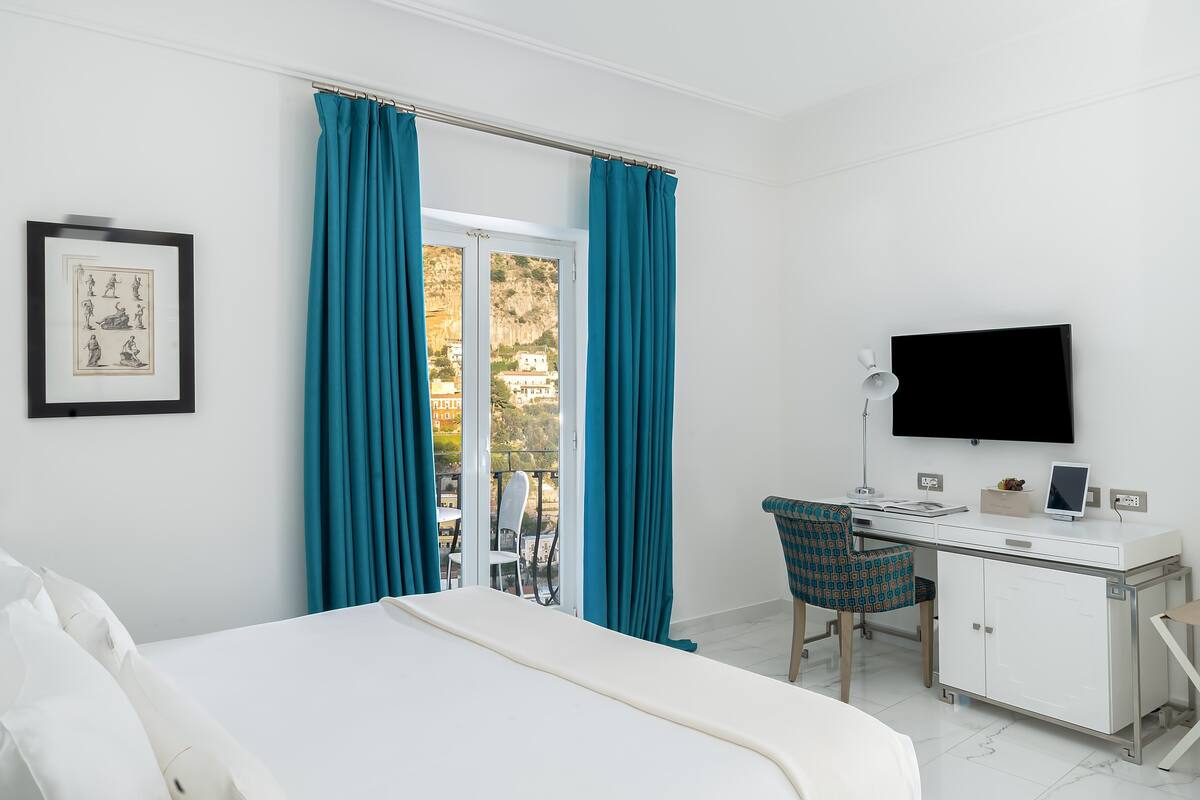 A hotel room with a bed, a desk with a computer, a TV, and a view of a balcony with blue curtains.