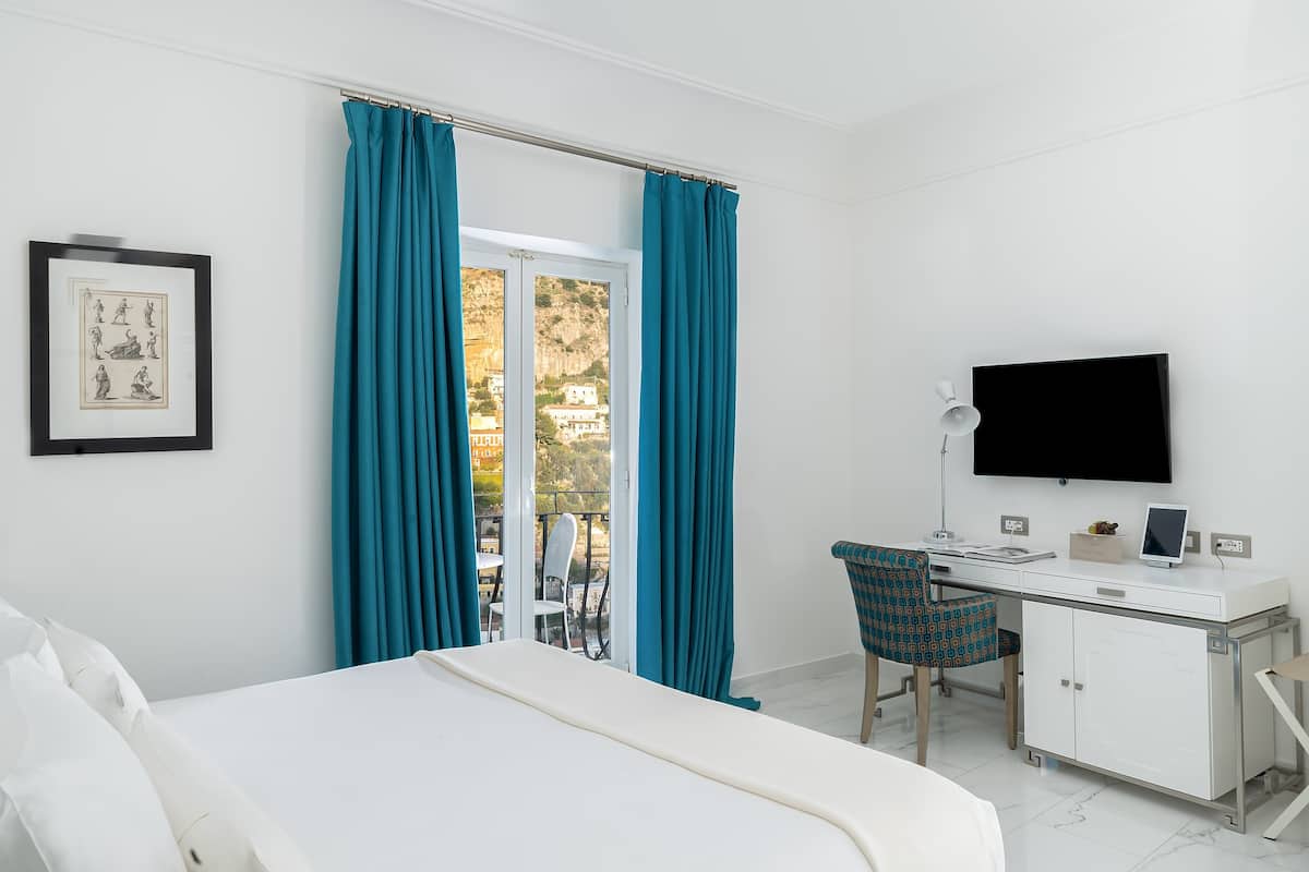 A hotel room with a bed, a desk with a computer, a TV, and a view of a balcony with blue curtains.